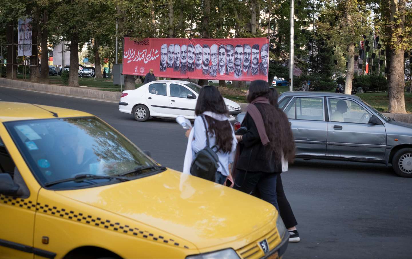 Young Iranian women walk past a banner featuring portraits of the IRGC generals and nuclear scientists who are killed in Israeli attacks, in northern Tehran, Iran, on June 30, 2025